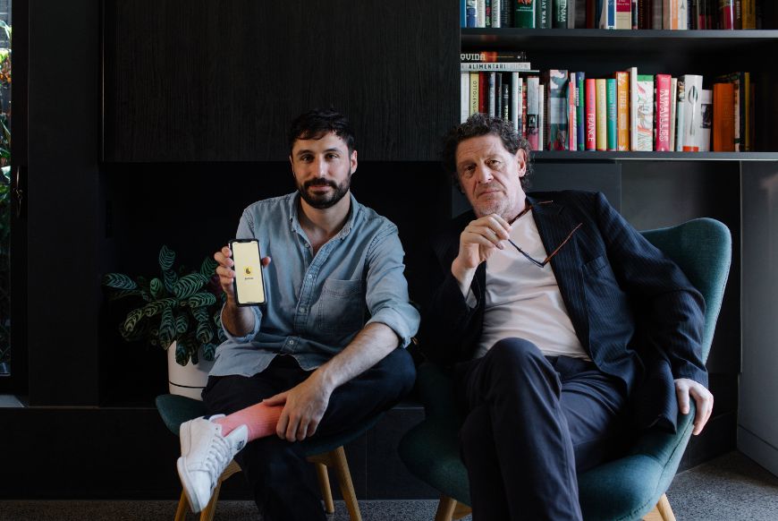 two men sit on chairs next to each other, one on the left is holding up a phone