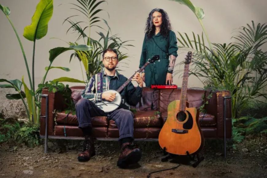 a man sitting on a sofa holding a guitar, and a woman standing behind him
