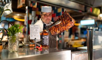 Chef behind the counter holding grilled meat towards the camera with tongs.