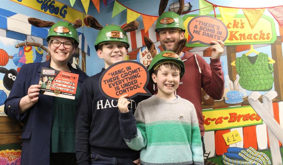 A family of four smiling in hard hats and holding Wallace & Grommit paper quotes and a completion certificate. In front of a colourful cartoon wall.