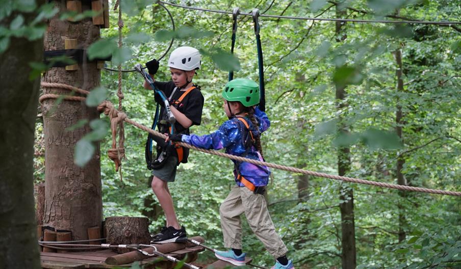 Two children on high ropes in the tree tops wearing helmets