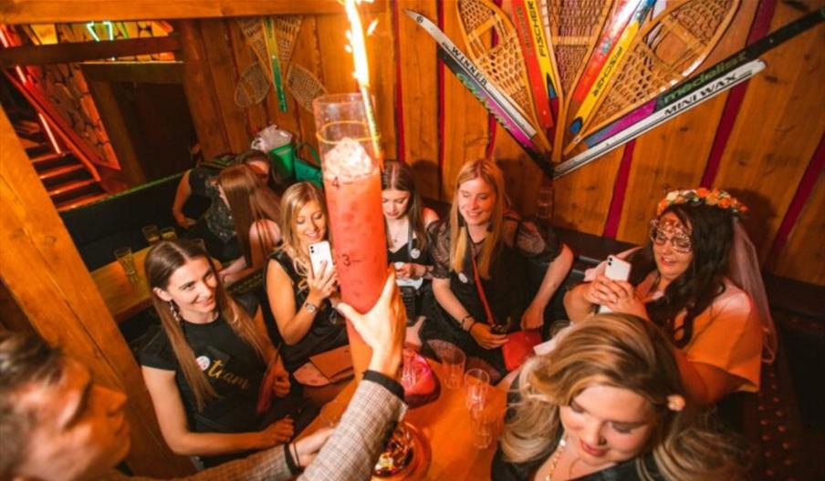 People around a table styled like a swiss chalet celebrating and being served food and drink