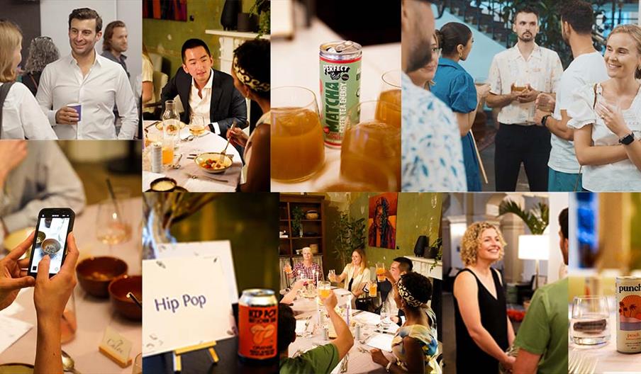 A lively room filled with people networking, chatting in small groups, and holding drinks. Tables display a variety of beverages, creating a warm and