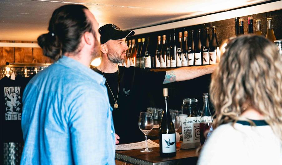 Adam at Saddle Goose, wearing a black t-shirt and cap, points at a row of wine bottles on a shelf while serving two customers.