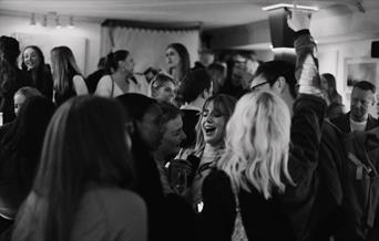 A black and white image of a room full of people, laughing and talking.