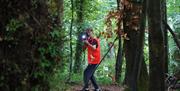 A teenage boy in an orange vest jacket pointing a camera with torch mounted on it within a forest environment.