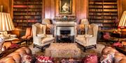 A wood panelled library sitting room with a chesterfield sofa and two cream sitting chairs around a small marble fireplace that has an old oil paintin