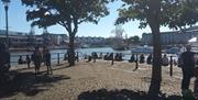 A view of Bristol's Harbourside, looking out across the water from next to Arnolfini building. People are sitting on the quayside enjoying the sunshin