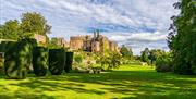 A view of green gardens with trees and box hedges with Berkeley Castle in the background