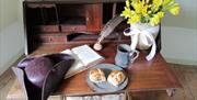 Work desk, with various items on it like a book, a quill and pot of ink, a hat, and items for breakfast