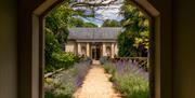 Looking through a stone archway down a gravel path lined with lavender that leads to a small stone building with a grey slate roof. It has a Cotswolds