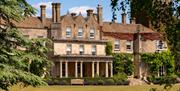 Stone front of a manor house with pillars, pitched roofs around windows, and tall chimney stacks. Blue sky in the background and grass and trees in th