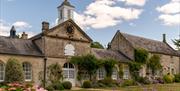 An old stone building with a clock in a pitched roof with a white turret behind it and a couple of outbuilding to the side. Several pretty arched wind