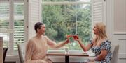 Two women sitting at a table by a window clinking their cocktail glasses together