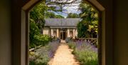 A stone archway leading down a lavender lined path to a sandstone outhouse with a slate pitched roof and wooden door. The entrance to the spa.