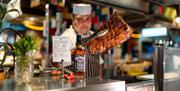 Chef behind the counter holding grilled meat towards the camera with tongs.