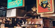 A female chef serving Indian food in a large serving counter with colourful signs and an ornate ceiling lampshade above