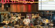 A chef serving up curries in large metal containers with a whole working kitchen in the background and large neon signs above his head saying 'curries