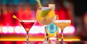 Three colourful cocktails in different shaped glasses sitting on a bar with neon lights behind them.
