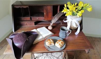 Work desk, with various items on it like a book, a quill and pot of ink, a hat, and items for breakfast