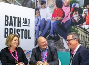 A woman and two men chatting in front of images of Bath and Bristol at World Travel Market