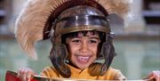 A young boy wearing a Roman helmet standing behind a shield by The Great Bath at The Roman Baths